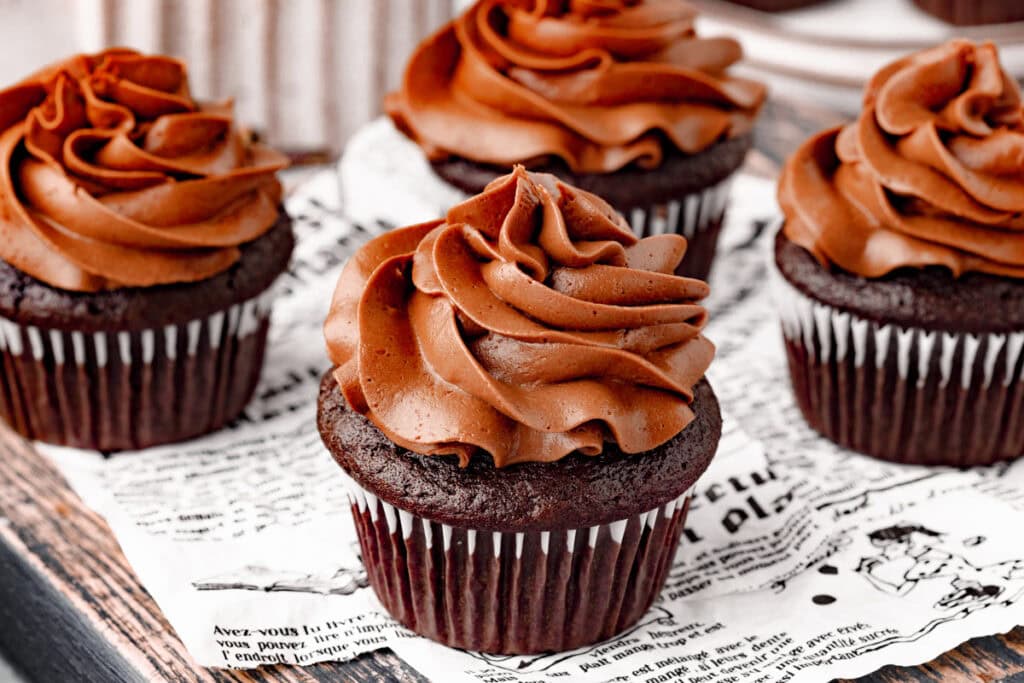 Four chocolate cupcakes with swirled chocolate frosting are arranged on a sheet of printed paper.