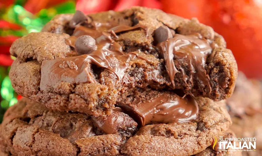 Close-up of stacked chocolate cookies with melted chocolate and chocolate chips, one cookie partially broken to show gooey center.