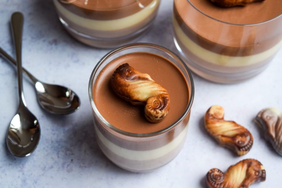 A glass jar with layered chocolate and vanilla dessert, topped with a chocolate-covered seahorse-shaped biscuit. Two spoons and similar biscuits are nearby.