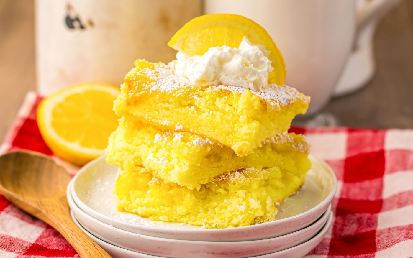 Three stacked lemon bars dusted with powdered sugar, topped with whipped cream and a lemon slice, on white plates next to a wooden spoon and a red checkered cloth.