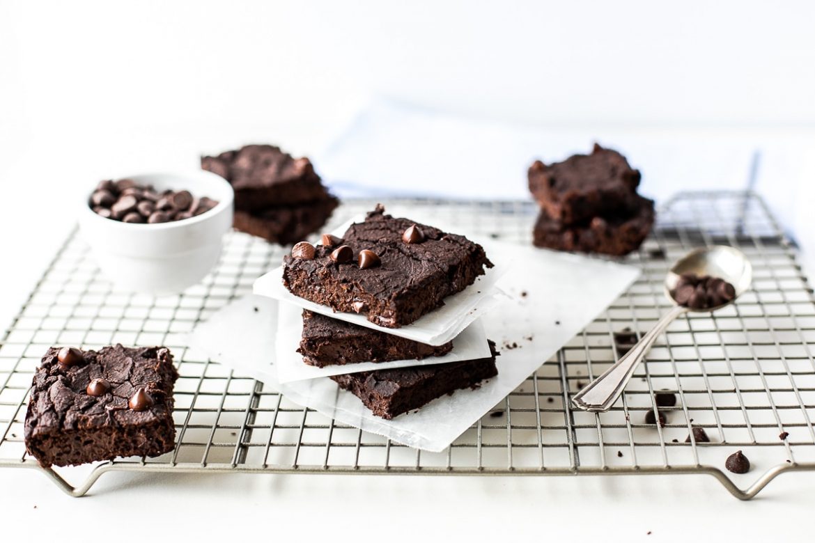 Brownies with chocolate chips are stacked on parchment paper atop a wire cooling rack, with a bowl of chocolate chips and a spoon nearby.