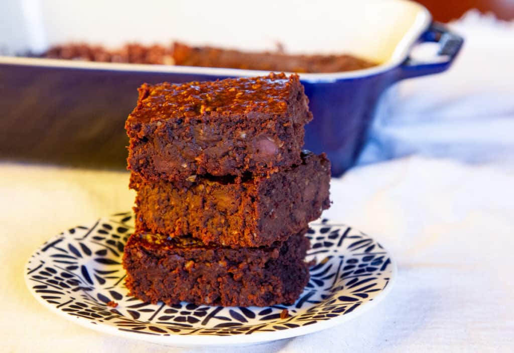 Three chocolate brownies stacked on a patterned plate, with a baking dish containing more brownies in the background.