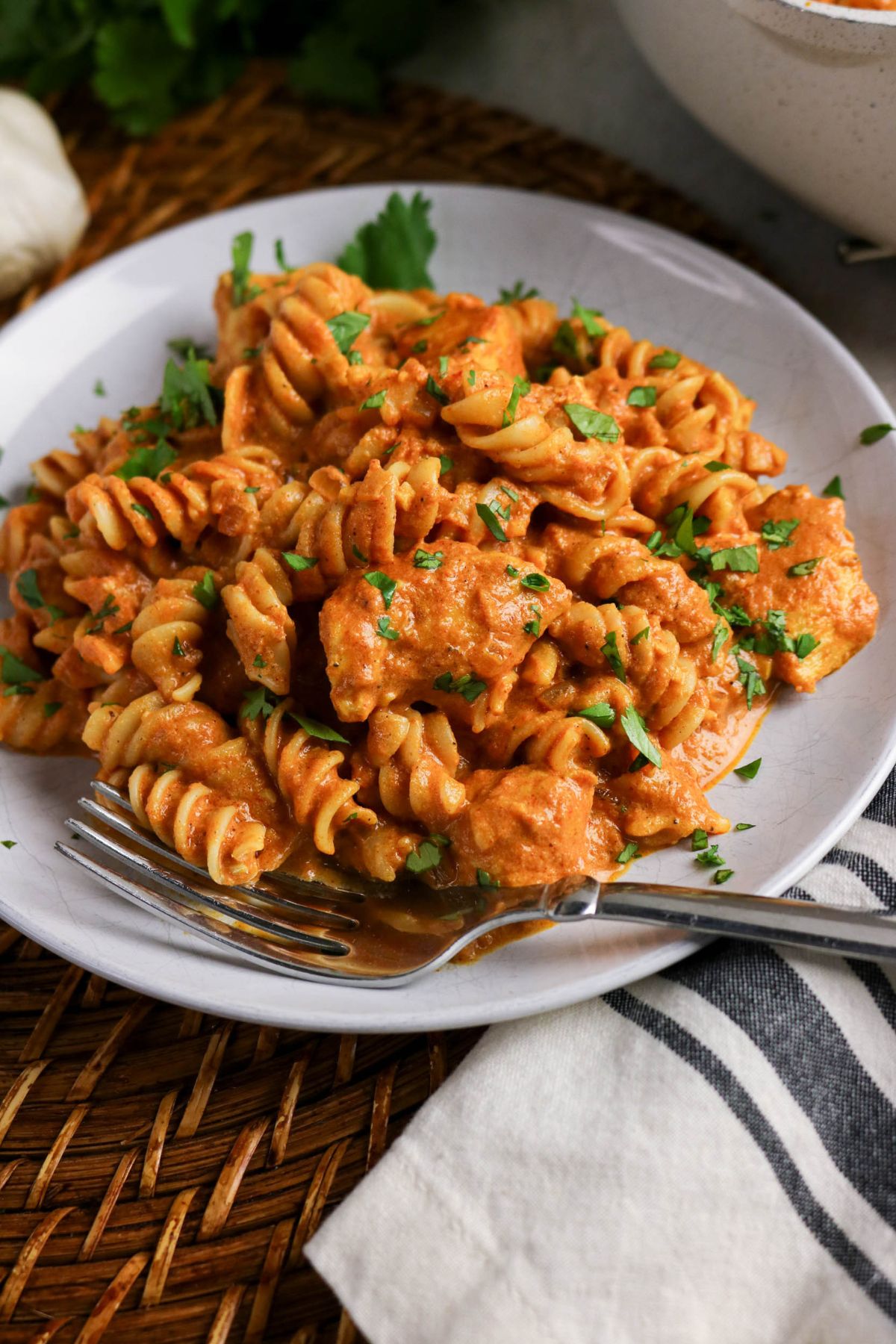 A plate of rotini pasta with pieces of chicken in a creamy tomato sauce, garnished with chopped herbs, with a fork on the side.