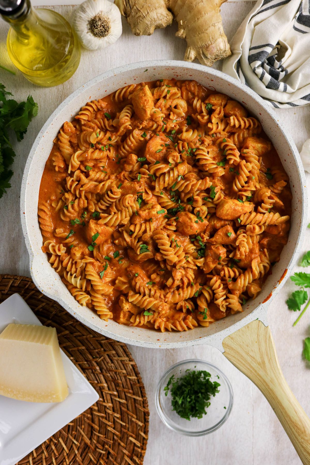 A skillet filled with rotini pasta and chunks of chicken in a creamy tomato sauce, garnished with herbs. Surrounding the skillet are garlic, ginger, oil, cheese, and fresh parsley.