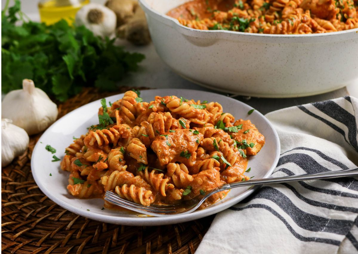 A white plate of rotini pasta with chunks of chicken in a creamy tomato sauce, garnished with chopped herbs, next to a fork, napkin, and fresh ingredients.