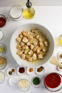 Overhead view of seasoned chicken pieces cooking in a pan, surrounded by small bowls of various spices, sauces, chopped herbs, and liquids on a white surface.