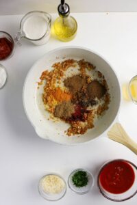 White skillet with sautéed onions and various ground spices. Surrounding the pan are bowls of tomato sauce, cream, herbs, garlic, oil, and other ingredients on a white surface.