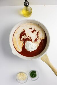 A white skillet with tomato sauce and cream is on a white surface, next to a bottle of olive oil, a small bowl of grated cheese, and a small bowl of chopped herbs.