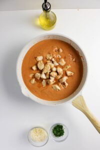 A white skillet with creamy tomato sauce and cooked chicken cubes, shown next to an olive oil bottle, grated cheese, and chopped herbs on a white surface.