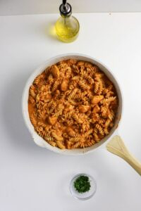 A white skillet with rotini pasta and sauce sits on a white surface next to a bottle of olive oil and a small bowl of chopped herbs.