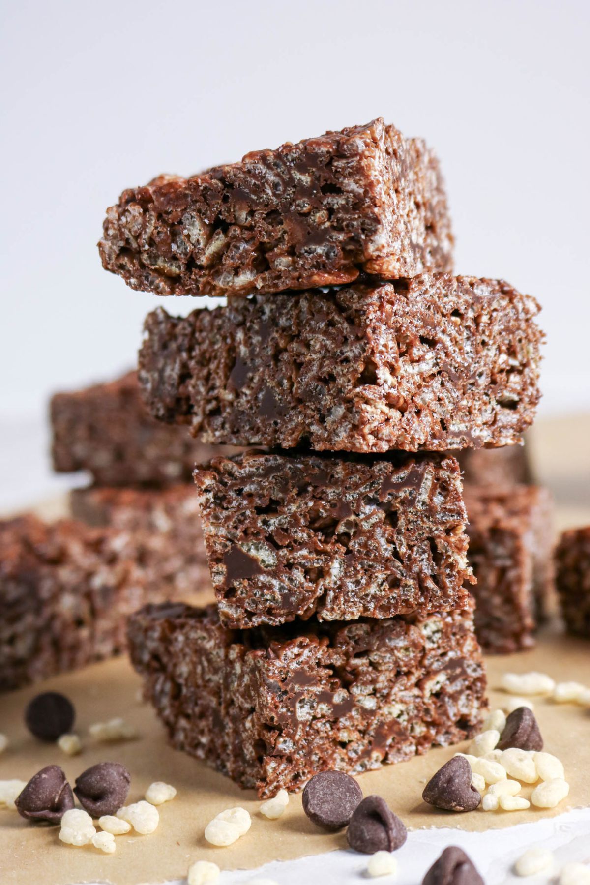 A stack of chocolate rice crispy treats on parchment paper, surrounded by chocolate chips and puffed rice cereal.