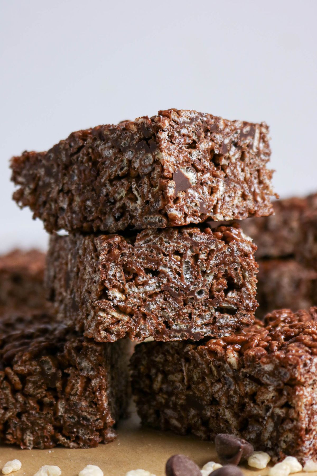 Three chocolate rice crispy treats are stacked on top of each other, with more treats and some chocolate chips and rice cereal pieces visible in the background.