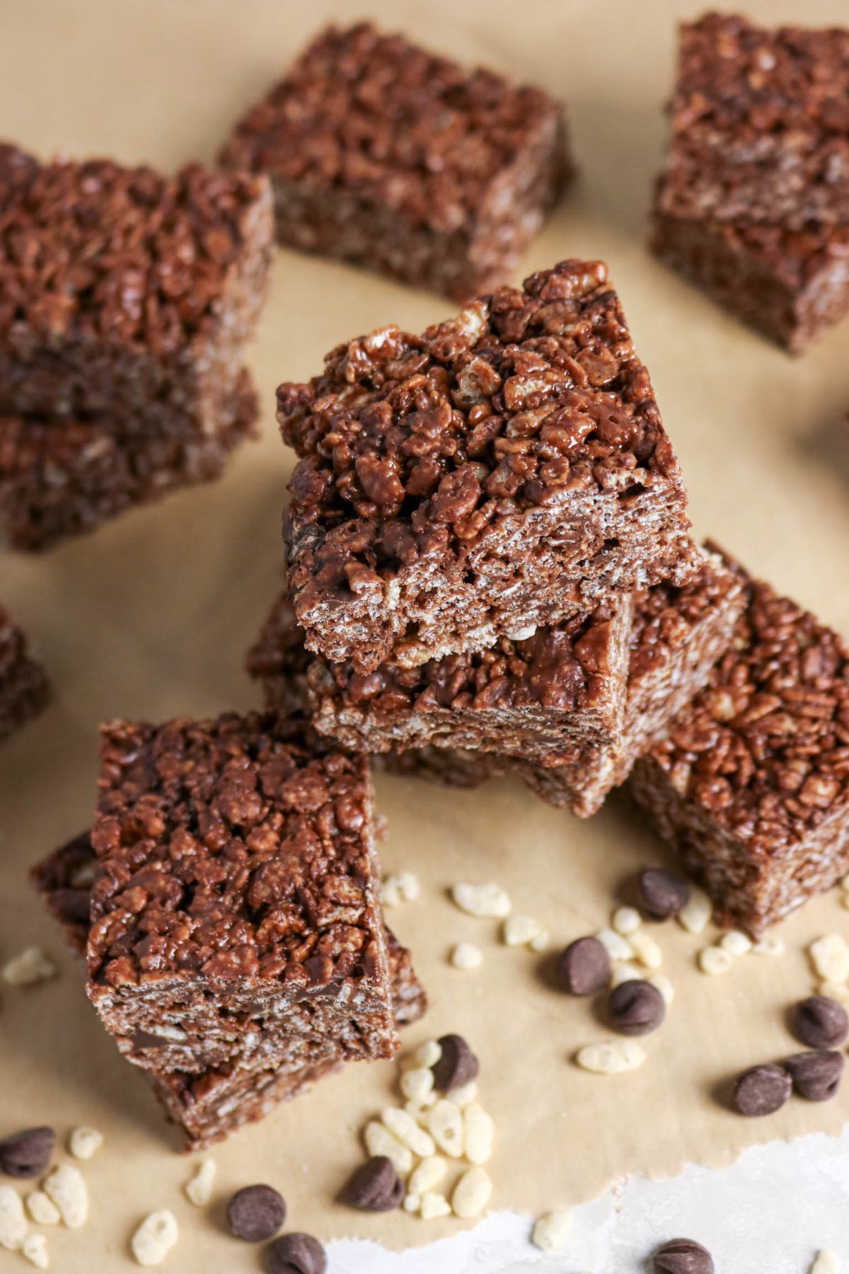 Chocolate rice crispy treats are stacked and arranged on parchment paper, with some chocolate chips and puffed rice scattered around.