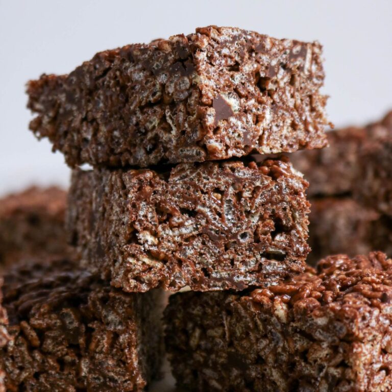 Three stacked chocolate rice crispy treats with a textured, crunchy appearance, surrounded by more treats in the background.