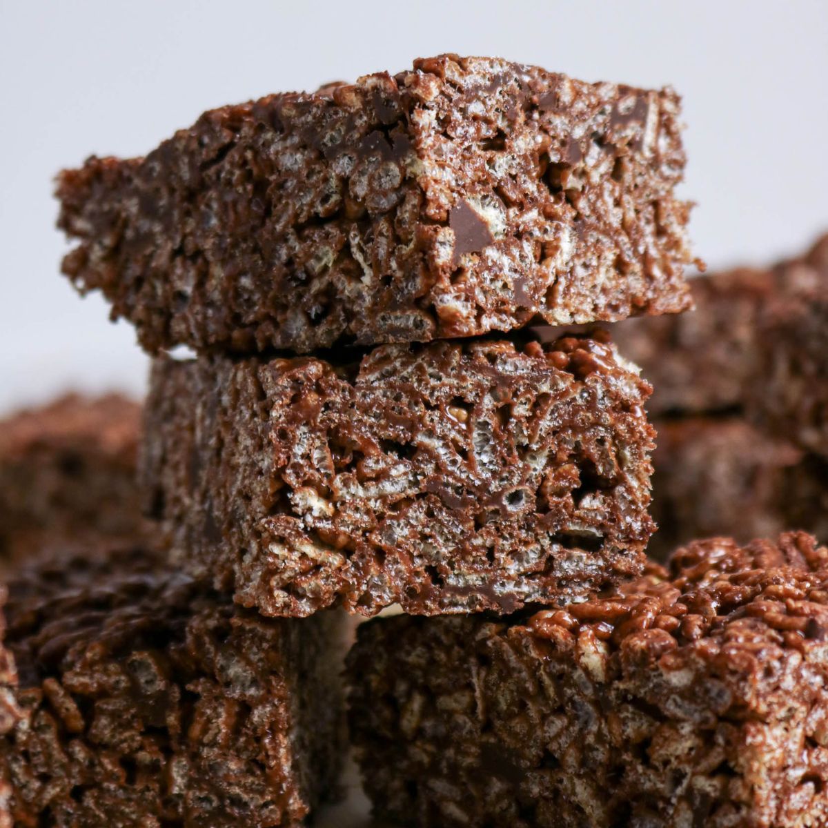 Three stacked chocolate rice crispy treats with a textured, crunchy appearance, surrounded by more treats in the background.