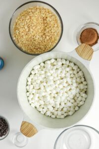 A bowl of mini marshmallows in a white pot next to bowls of crispy rice cereal, cocoa powder, and other ingredients on a white surface.