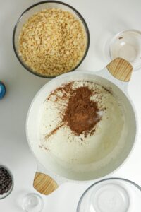 A mixing bowl with cream and cocoa powder, surrounded by bowls of chopped nuts, chocolate chips, and empty glass bowls on a white surface.