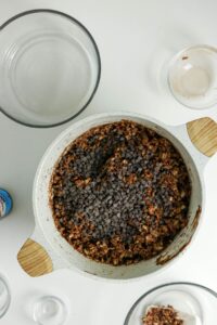 A white mixing bowl filled with oatmeal cookie dough topped with chocolate chips, surrounded by empty glass bowls on a white surface.