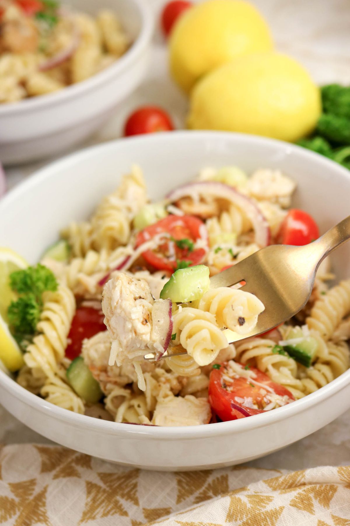 A fork lifts a bite of pasta salad with rotini, chicken, cucumbers, tomatoes, red onion, and parsley from a white bowl. Lemons and vegetables are in the background.
