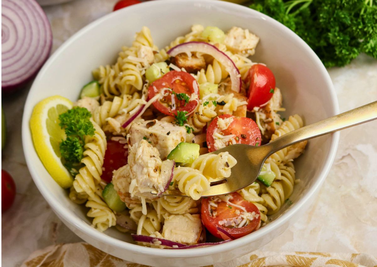 A white bowl filled with rotini pasta salad, cherry tomatoes, cucumber, red onion, chicken pieces, grated cheese, and a parsley and lemon garnish. A fork is placed inside the bowl.