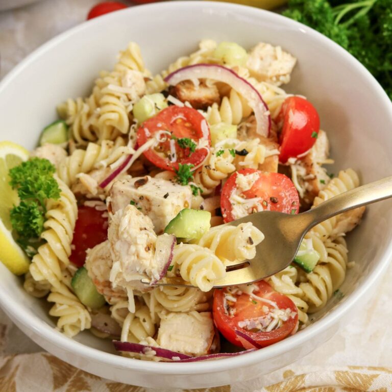 A bowl of rotini pasta salad with chunks of chicken, cherry tomatoes, cucumber, red onion, shredded cheese, and a parsley garnish. A lemon slice and parsley are on the rim; a fork rests inside.