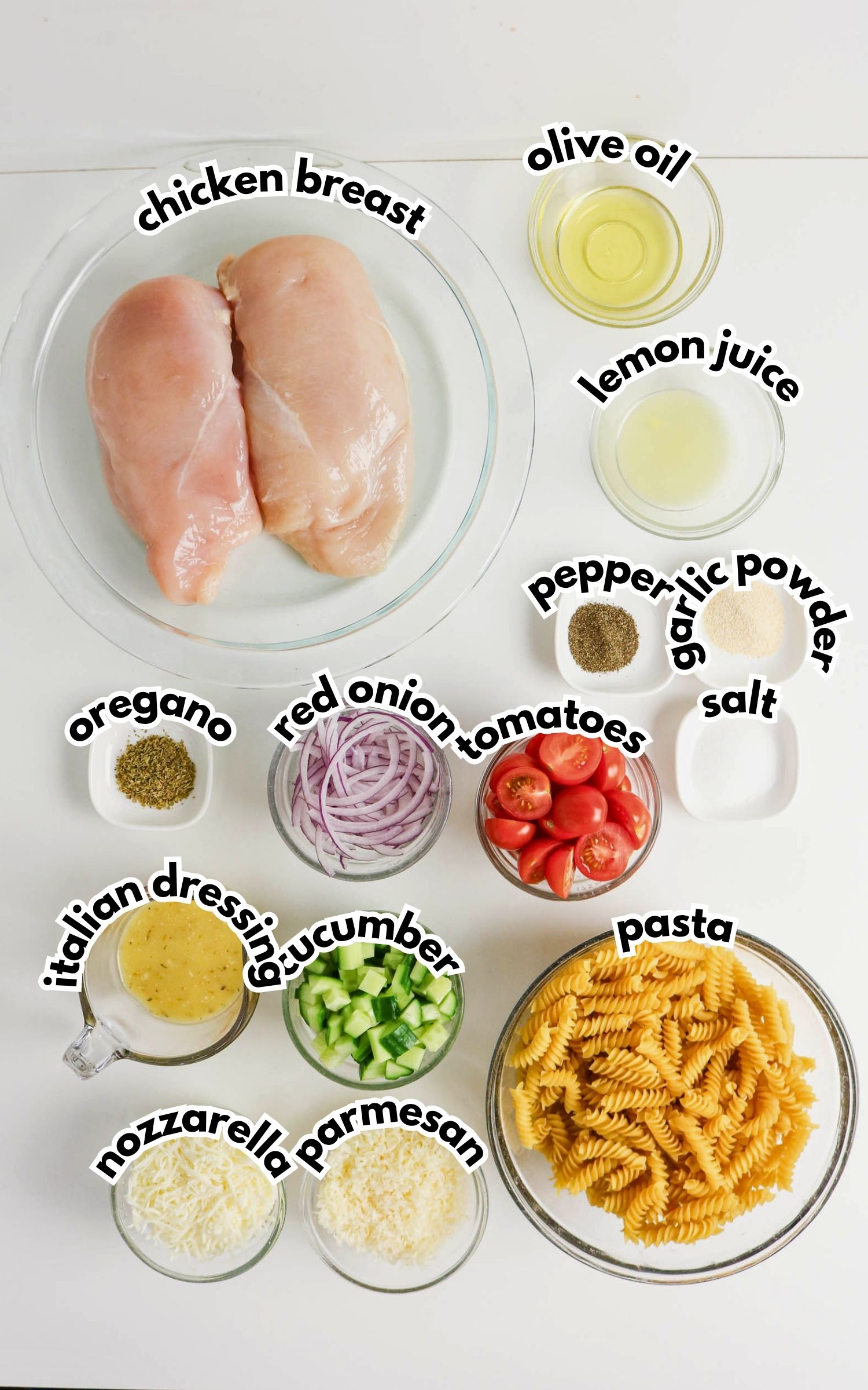 Ingredients for a pasta salad arranged on a white surface, including chicken breast, pasta, vegetables, shredded cheese, and various seasonings in separate bowls.