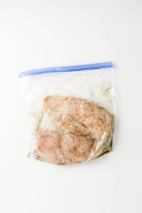Raw chicken breasts with seasoning inside a sealed plastic zip-top bag, placed on a white surface.