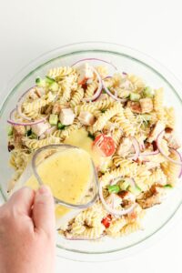 A hand pours creamy dressing over a bowl of rotini pasta salad with grilled chicken, cherry tomatoes, red onion, and cucumber.