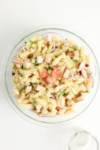 A glass bowl filled with rotini pasta salad, featuring pieces of chicken, cherry tomatoes, sliced red onion, cucumber, and a light dressing.