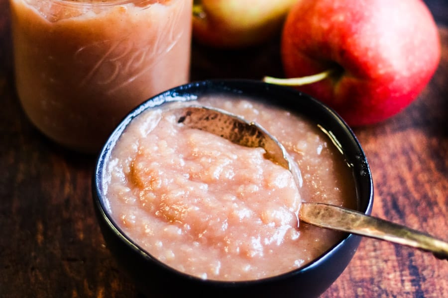A bowl of applesauce with a spoon, a glass jar of applesauce, and two red apples on a wooden surface.