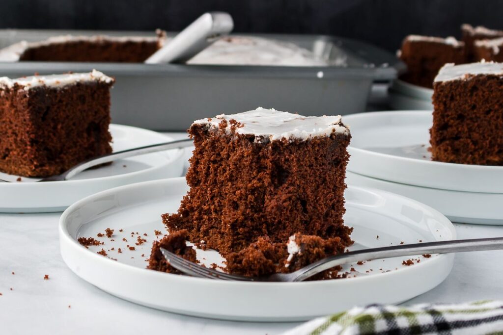 A square slice of chocolate cake with white icing on a white plate and fork, with more slices and a baking pan in the background.