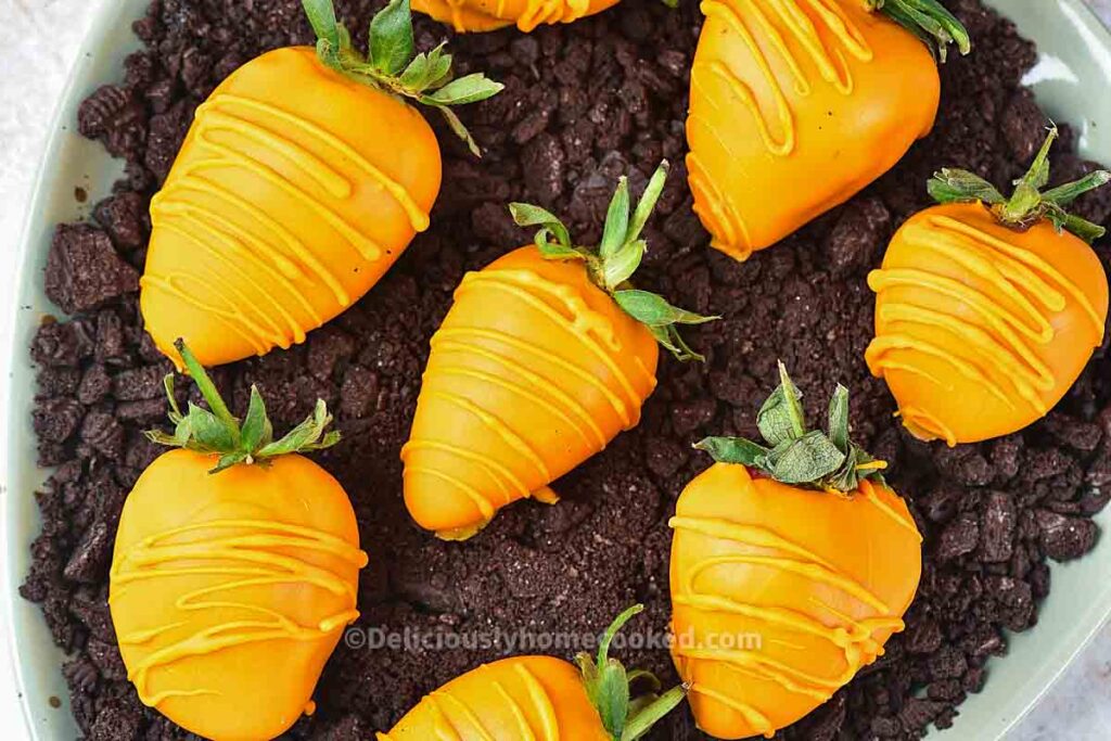 Seven strawberries coated in orange chocolate are arranged on a plate of crushed chocolate cookies, resembling carrots in soil.
