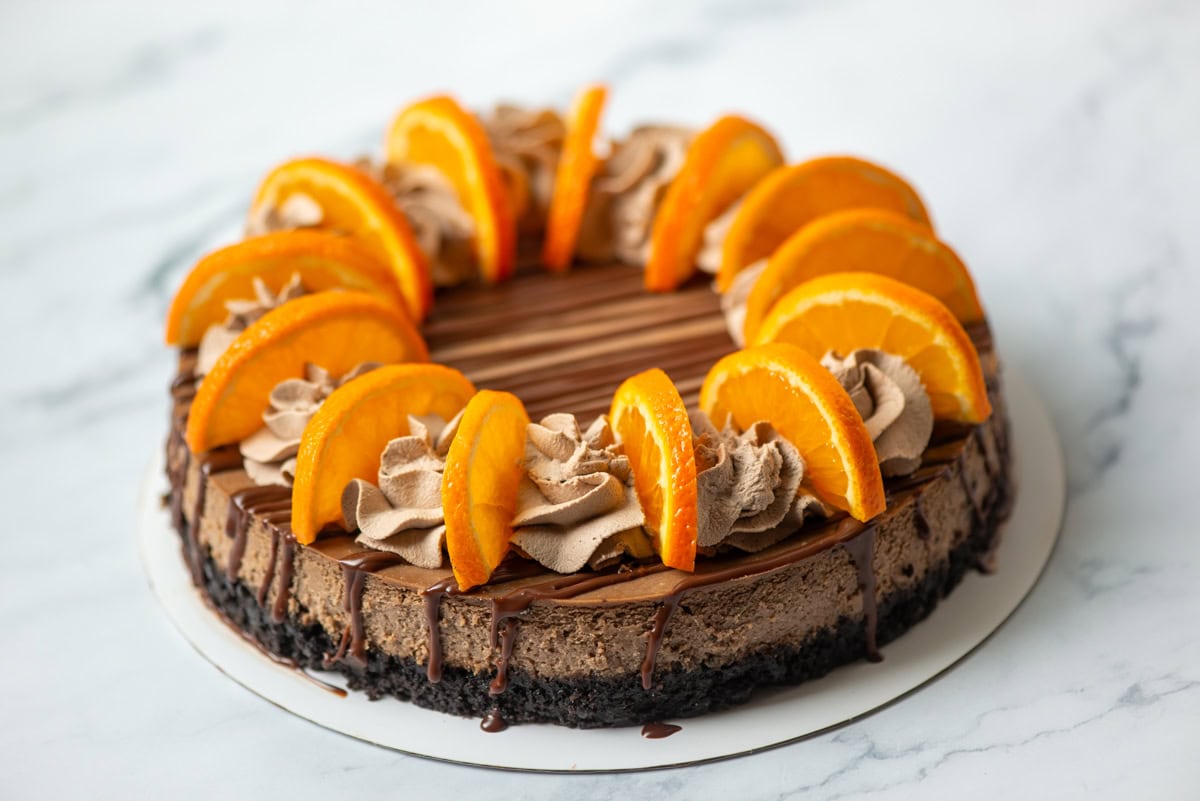 A chocolate cheesecake topped with chocolate cream swirls and orange slices, displayed on a white round base against a marble background.