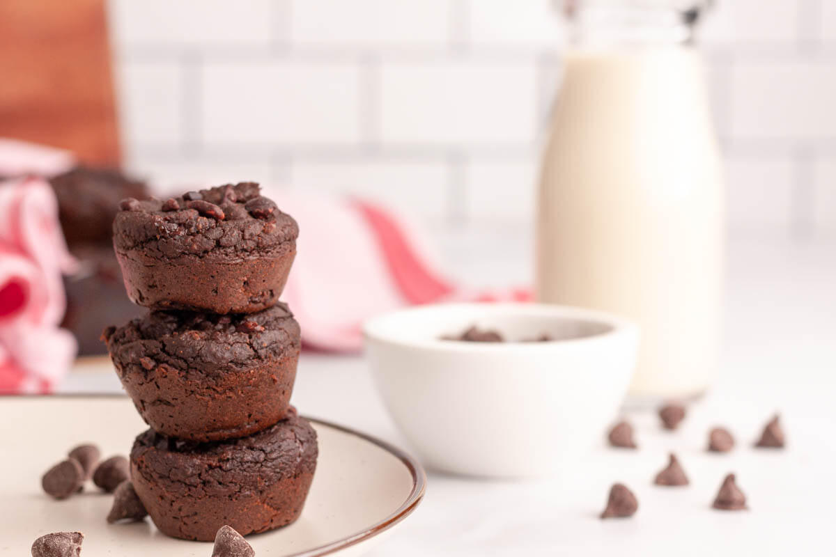 Three chocolate brownies stacked on a plate beside chocolate chips, a bowl, and a bottle of milk, with a pink cloth and tiled background.