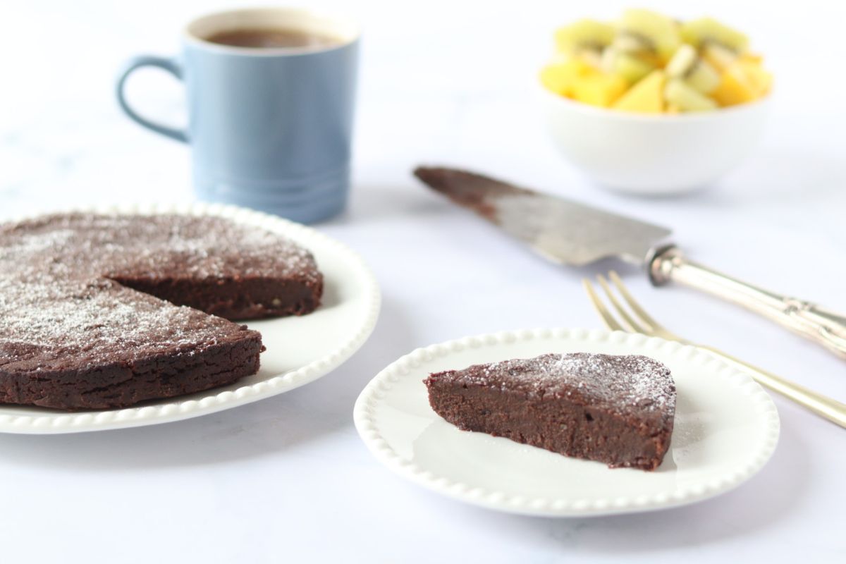 A slice of chocolate cake on a plate, with the rest of the cake, a cup of coffee, a fork, a cake server, and a bowl of cut fruit in the background.