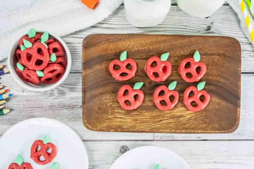 Red pretzels shaped and decorated to resemble apples with green leaves, arranged on a wooden tray and plates, with pencils and milk bottles nearby.