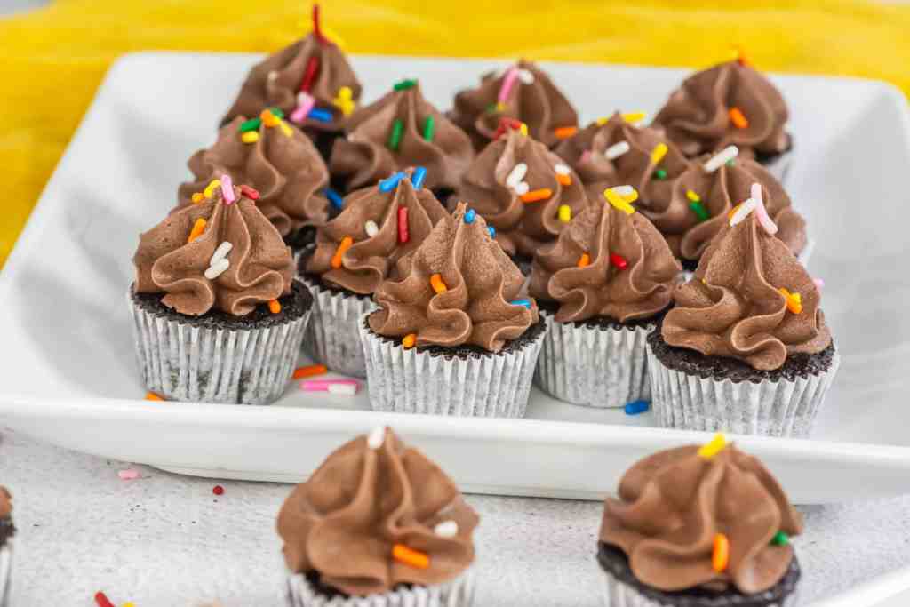 A group of chocolate cupcakes with chocolate frosting and colorful sprinkles arranged on a white rectangular platter.