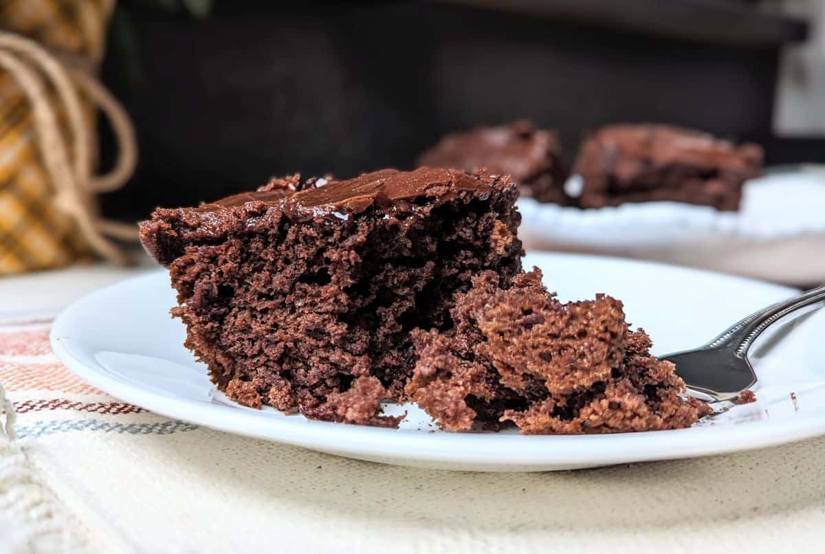 A slice of chocolate cake on a white plate with a fork holding a piece of the cake; background shows more cake pieces out of focus.