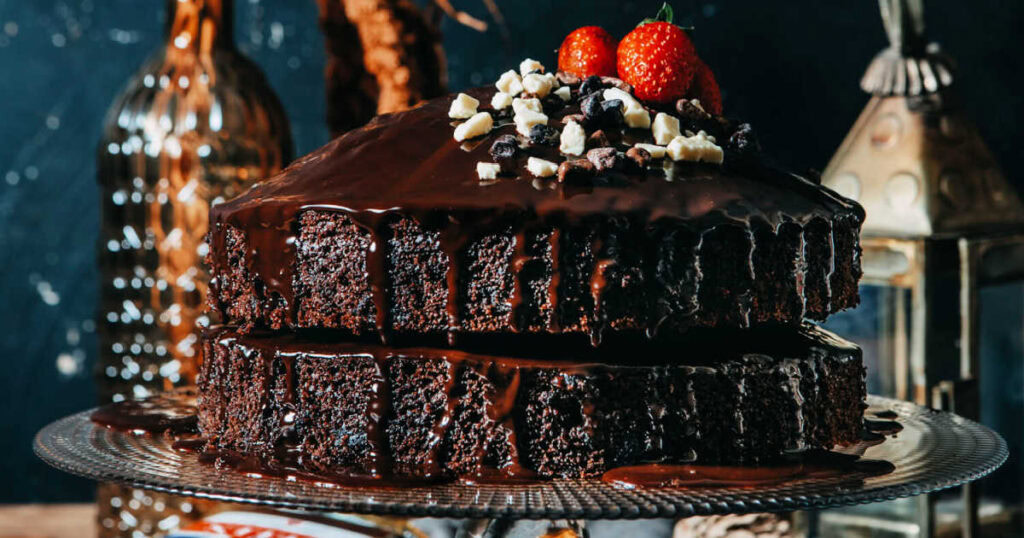Two layers of chocolate cake covered in chocolate ganache, topped with chopped nuts, chocolate pieces, and strawberries, displayed on a glass plate.