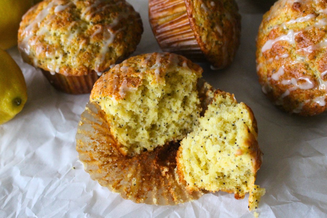 A close-up of lemon poppy seed muffins with glaze on top, one muffin split in half to show its moist texture, on a white parchment background.