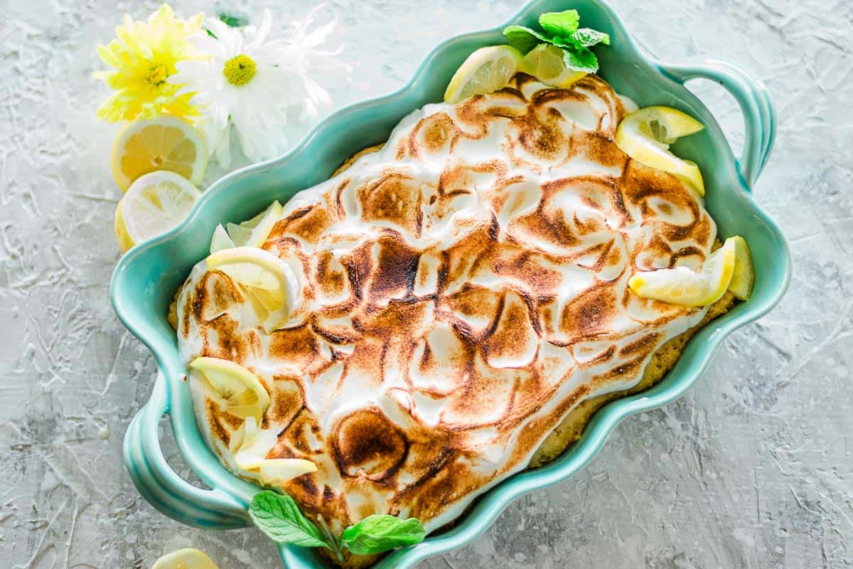 A baked lemon meringue dessert in a turquoise dish, garnished with lemon slices and mint, sits on a textured surface with flowers and lemon slices nearby.