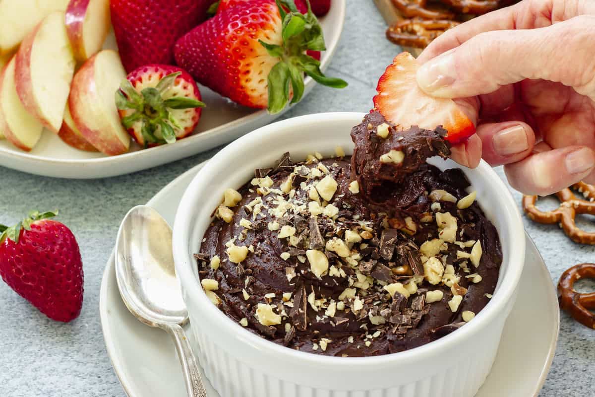 A hand dips a strawberry into a bowl of chocolate dessert topped with chopped nuts. Sliced apples, whole strawberries, and pretzels are on a plate nearby.