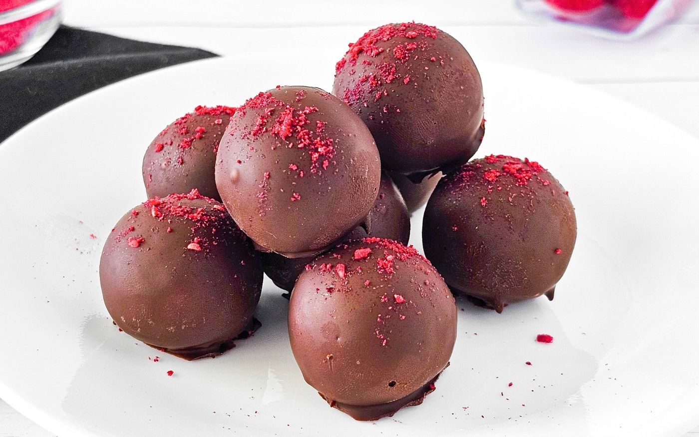 A white plate with seven chocolate truffles, each topped with a sprinkle of red powder, arranged in a small pile.