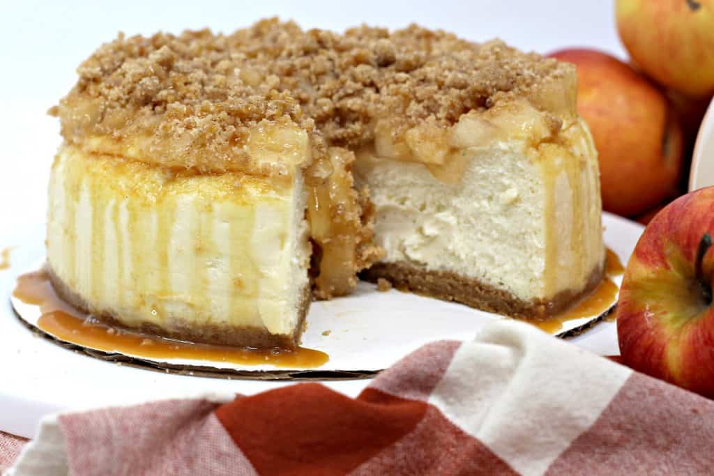 A cheesecake with a crumb topping and caramel drizzle sits on a white surface, with a slice removed. Apples and a red-and-white cloth are nearby.