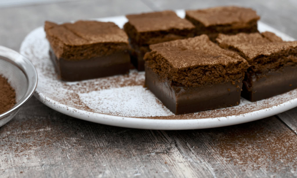 A white plate holds six pieces of chocolate layered cake dusted with cocoa powder, placed on a rustic wooden surface.