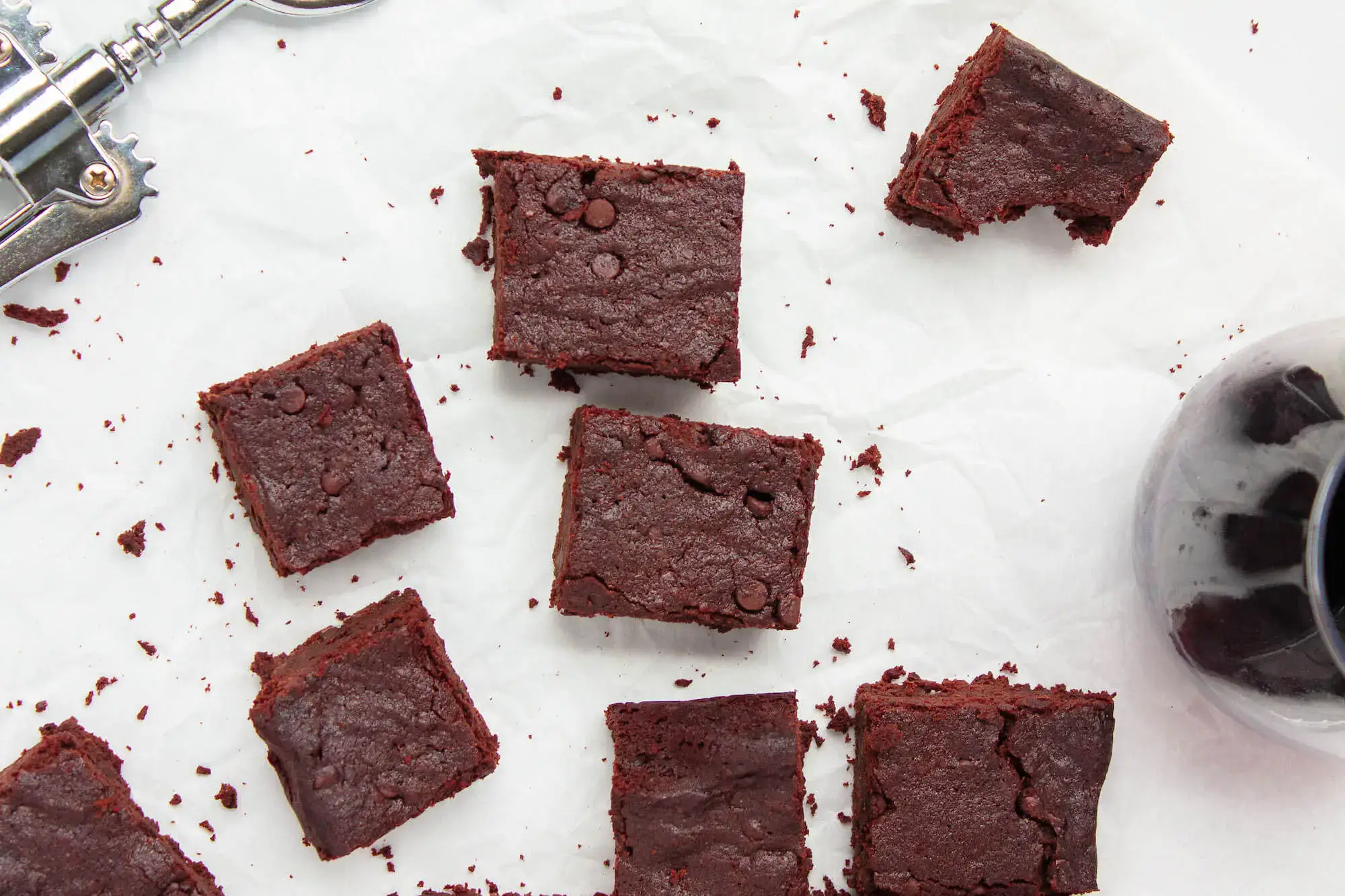Several square pieces of chocolate brownies are arranged on white parchment paper, with a corkscrew and a glass of red wine visible nearby.