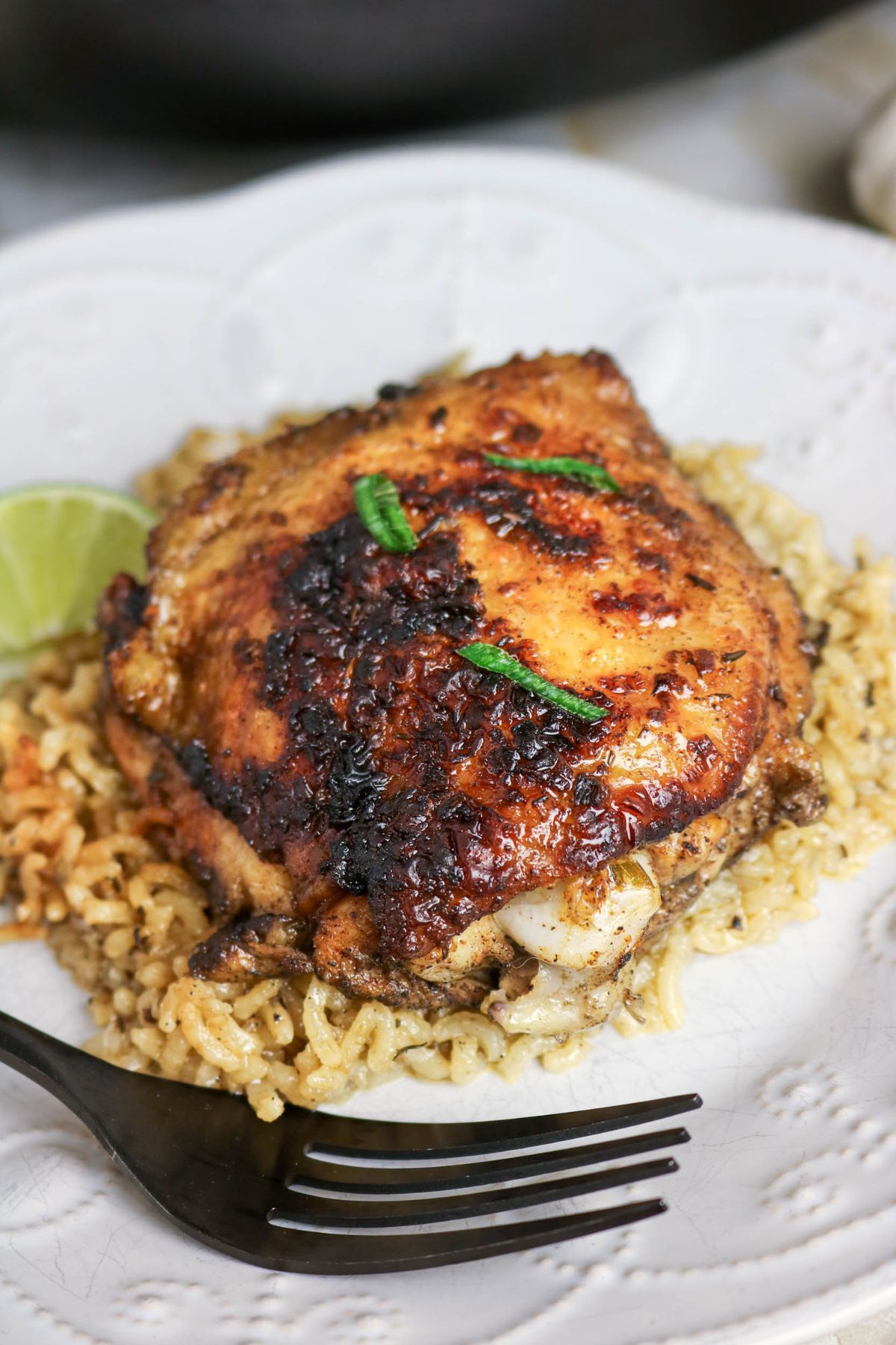 A piece of seasoned, grilled chicken thigh served on a bed of rice with a wedge of lime and garnished with chopped green herbs on a white plate. A fork is placed beside the food.
