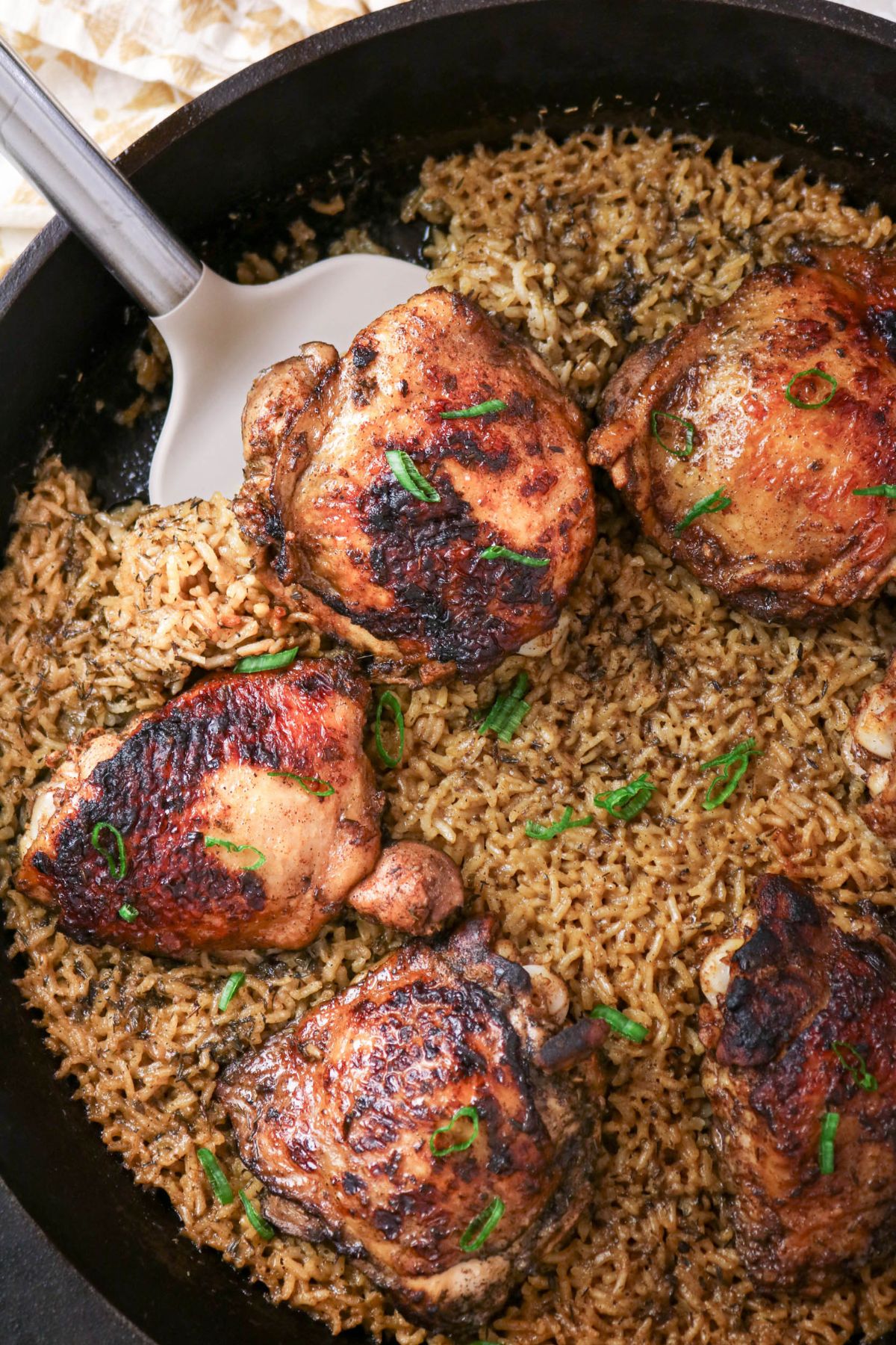 A skillet filled with browned chicken thighs and seasoned rice, garnished with chopped green onions, with a spatula resting in the pan.