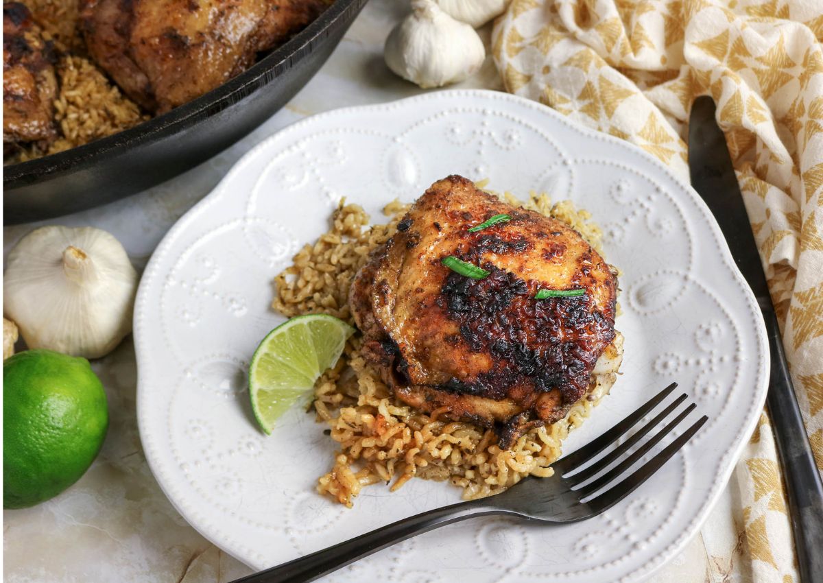 A piece of seasoned grilled chicken on a bed of rice, garnished with green herbs and served with a lime wedge on a white plate. A fork and knife are placed beside the plate.