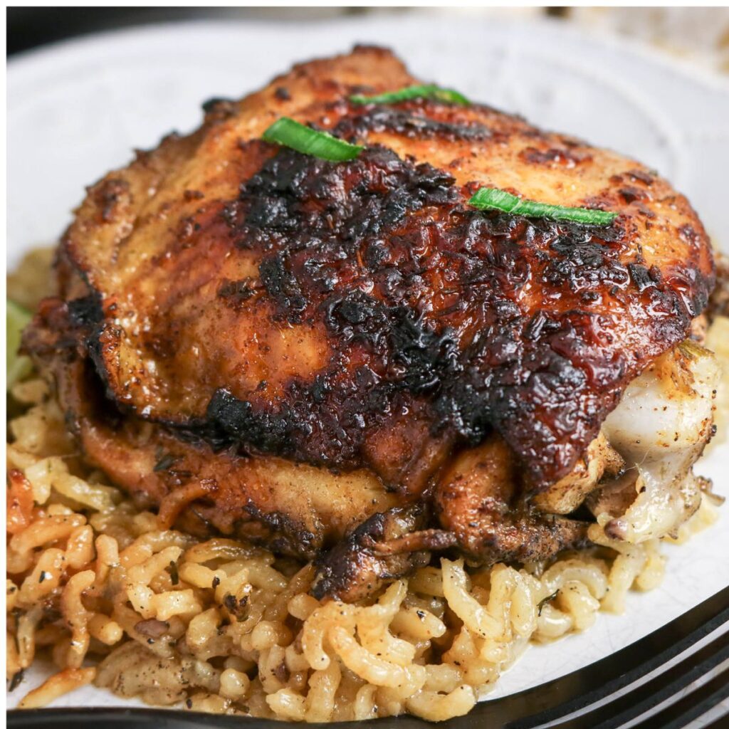 Close-up of a seasoned, browned chicken thigh served on a bed of cooked rice on a white plate.
